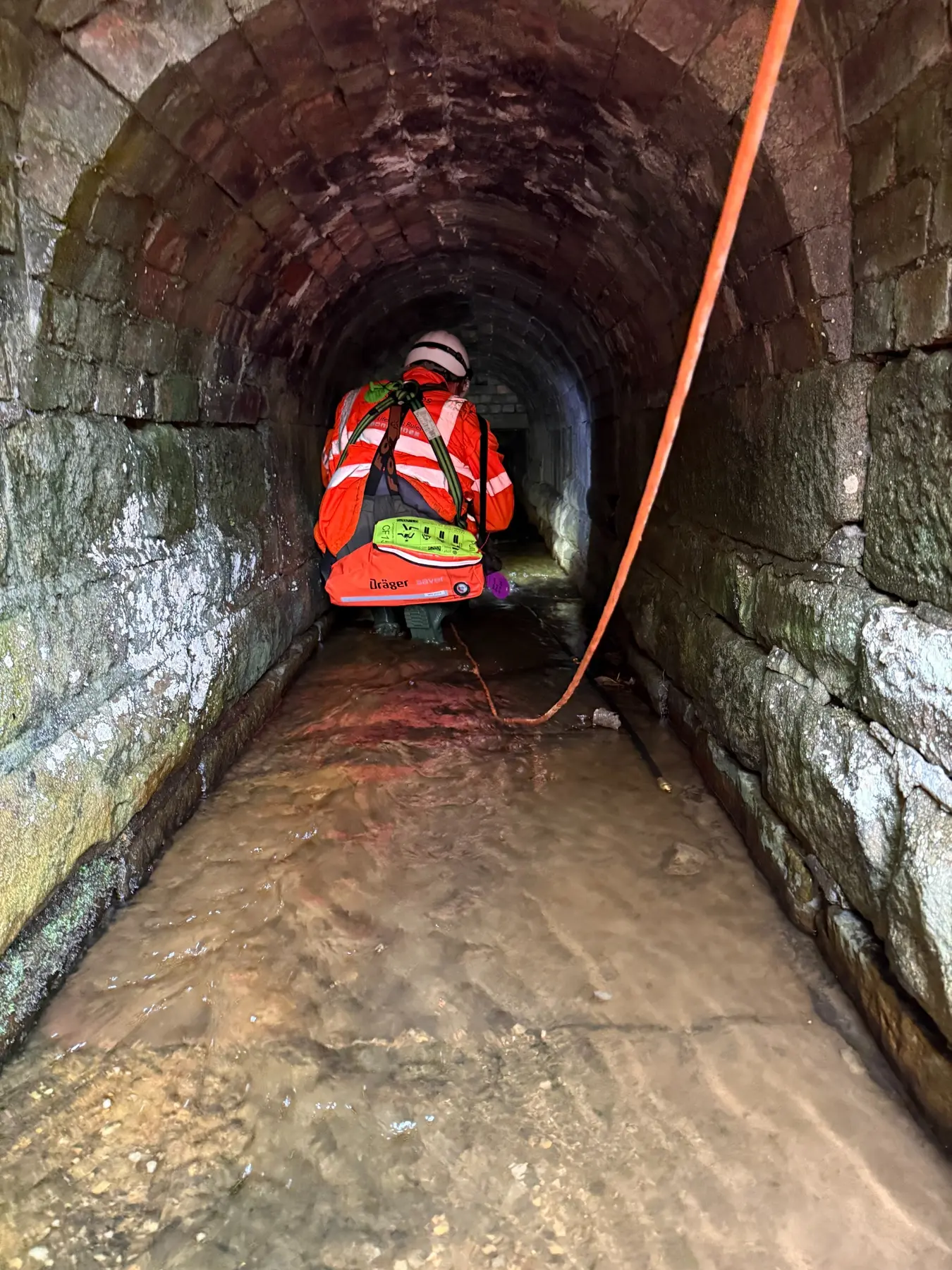 Confined-space operative entering a narrow water-filled masonry culvert with communications line